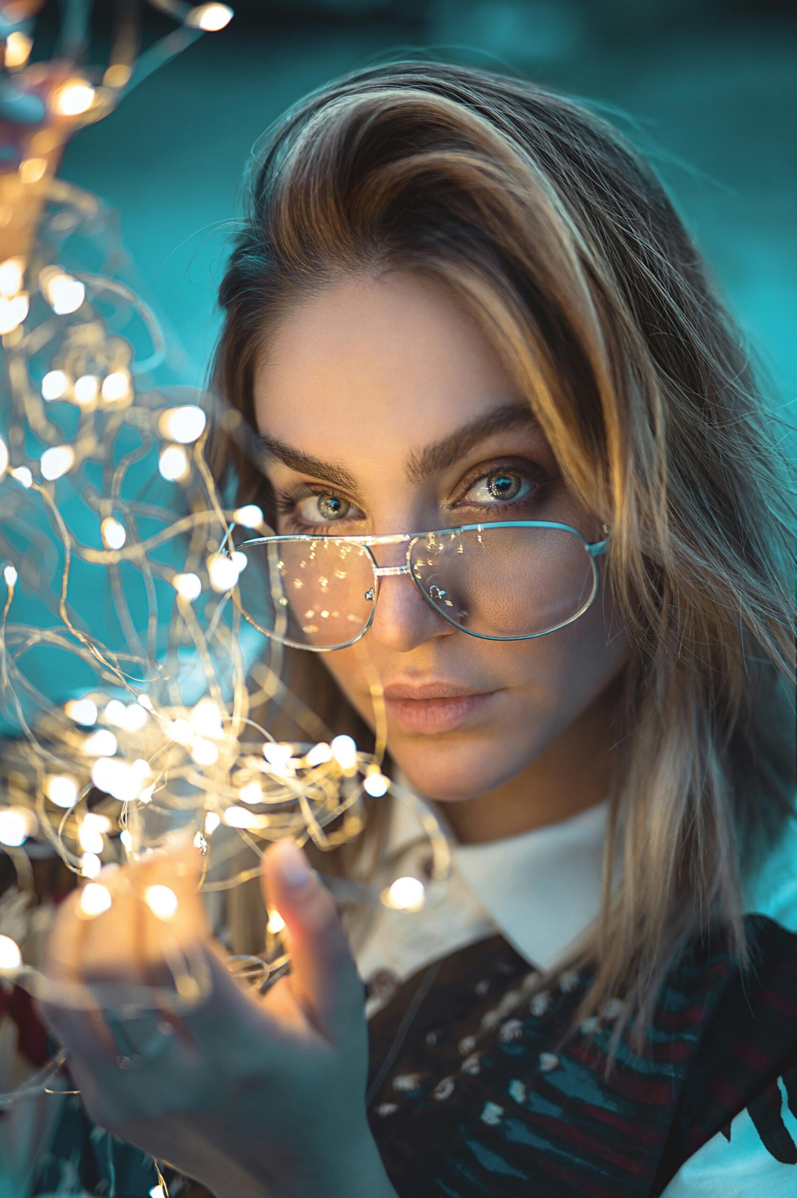 Portrait of a stylish woman with glasses holding string lights, creating a bokeh effect.